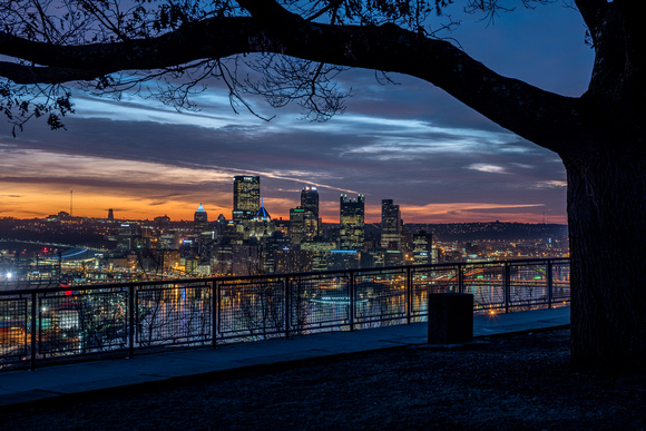 A tree frames Pittsburgh from the West End uner colorful skies