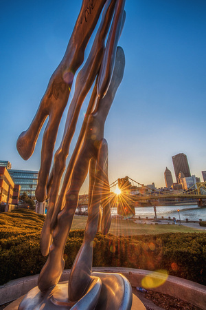 The sun rises beside a sculpture on the North Shore of Pittsburgh