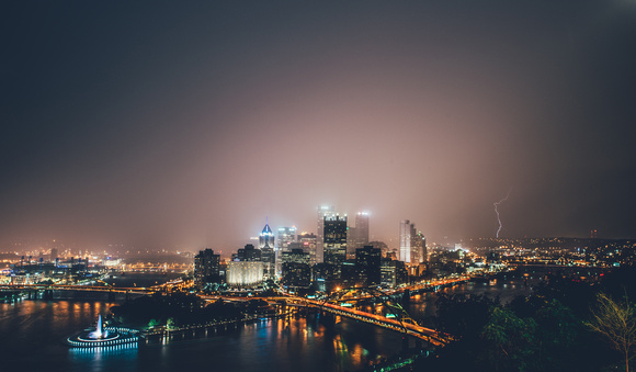The rain is illuminated by the lights of Pittsburgh as lightning strike sin the background