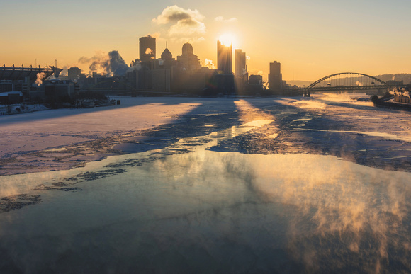 The sun sits on on the top of the Pittsburgh skyline over an icy Ohio River